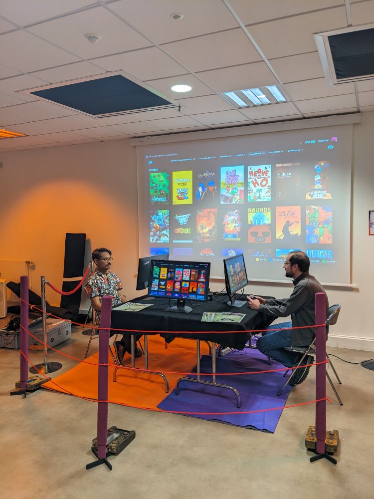 2 joueurs face à face autour d'une table, assis manettes en main face à leurs écrans respectifs. Au premier plan : un décor de ring de box. En fond : une sélection de jeux, projetée au mur.