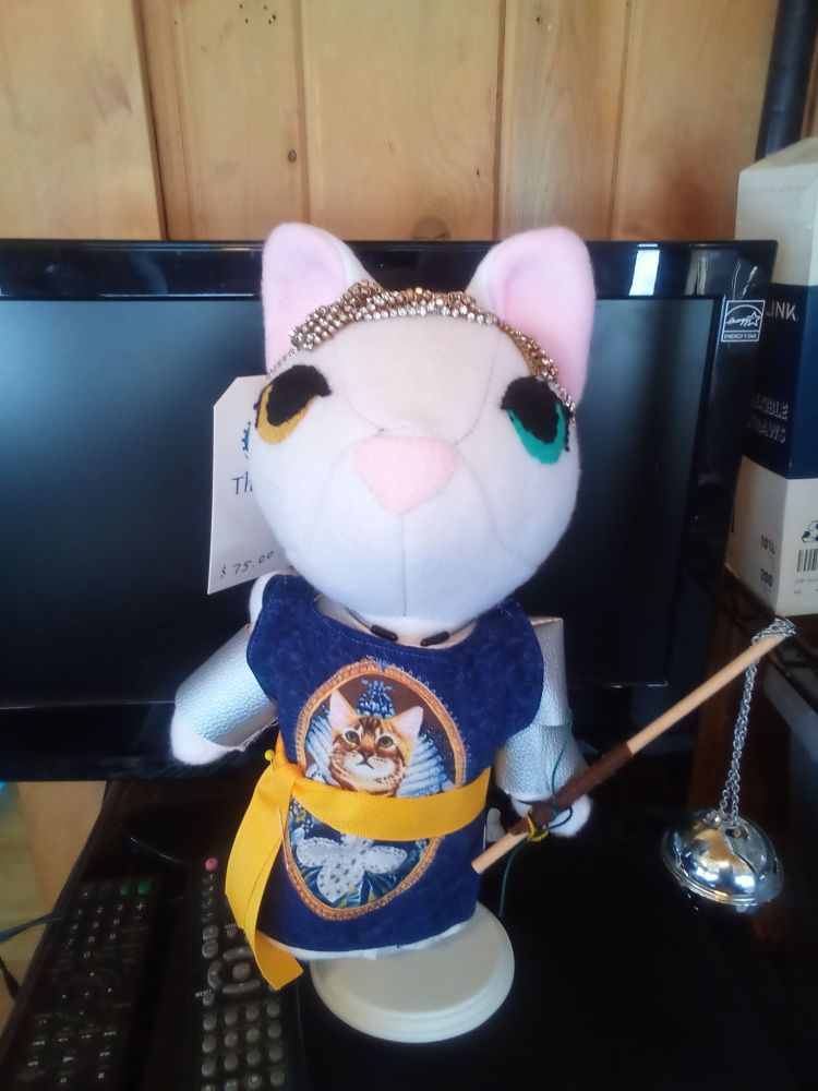 Anthropomorphic stuffed animal, white cat with one blue eye and one yellow eye, dressed in plate mail armor with a blue tabard. The tabard has a picture of Queen Elizabeth I as a cat on it. The cat holds a flail with a head made of a large jingle bell.