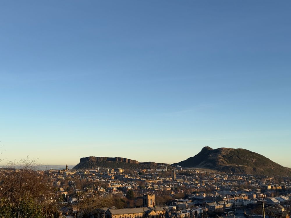 A view of Edinburgh from Blackford Hill. In the foreground are streets of tenement flats, houses and other buildings. In the distance you see the Salisbury Crags on the left, with Arthur’s Seat on the right. A clear, blue sky lies above as the sun rises in the morning. 