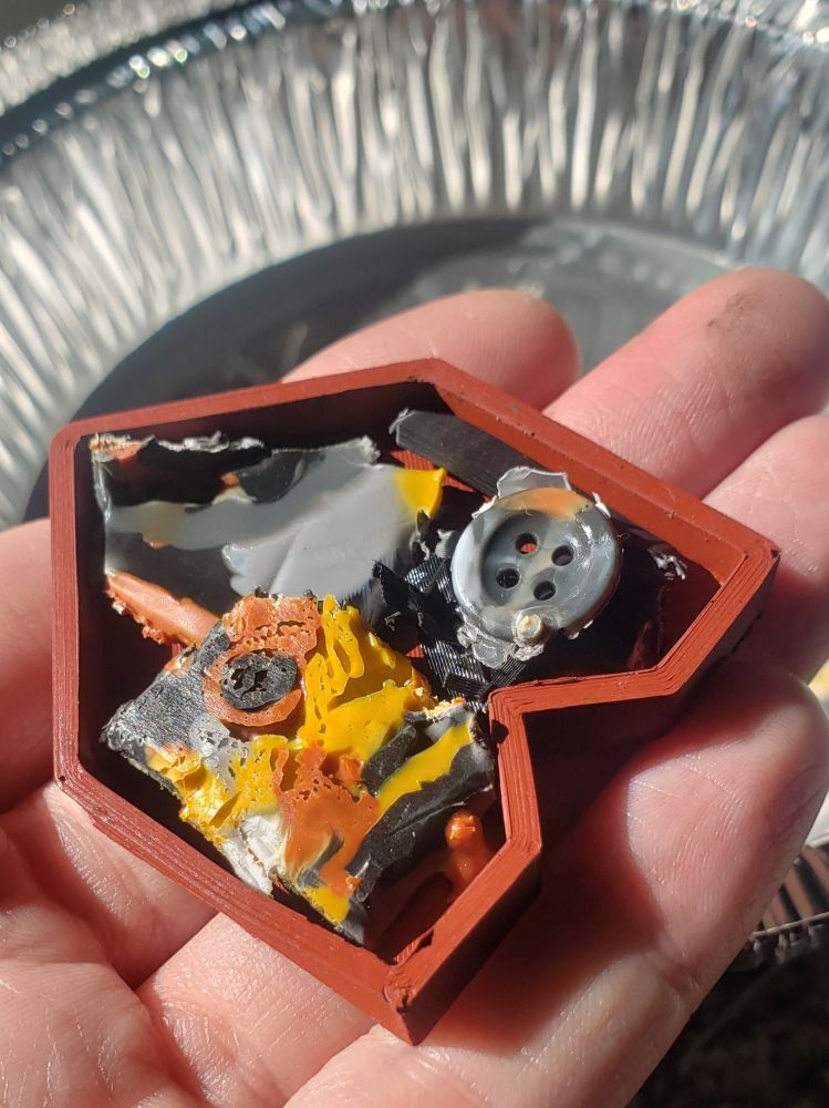 A hand holding a brick-red rubber mould filled with irregular chunks of plastic, one of which looks like a grey shirt button.