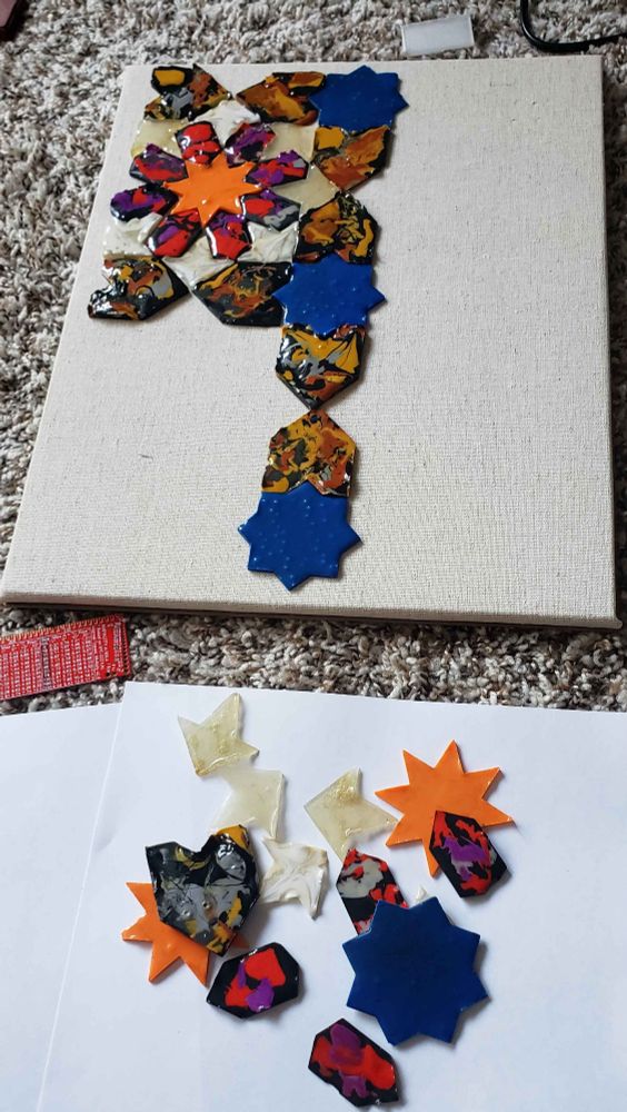 A photo of a canvas board partly covered in zellij tiles. The 8-pointed star pieces are solid blue or solid orange color, and the rest of the tiles are messy tie-dye colors. More tiles sit on a piece of paper next to the canvas, waiting to be placed.