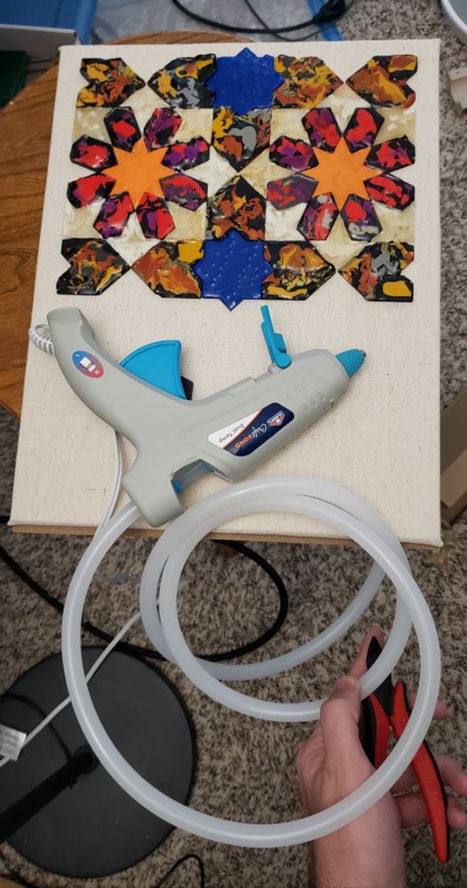 The canvas from the previous post, now fully tiled on the top half. A hot glue gun is lying on the empty bottom half. A 5-foot-long continuous coil of hot glue stick is spooling out the back of the glue gun.
