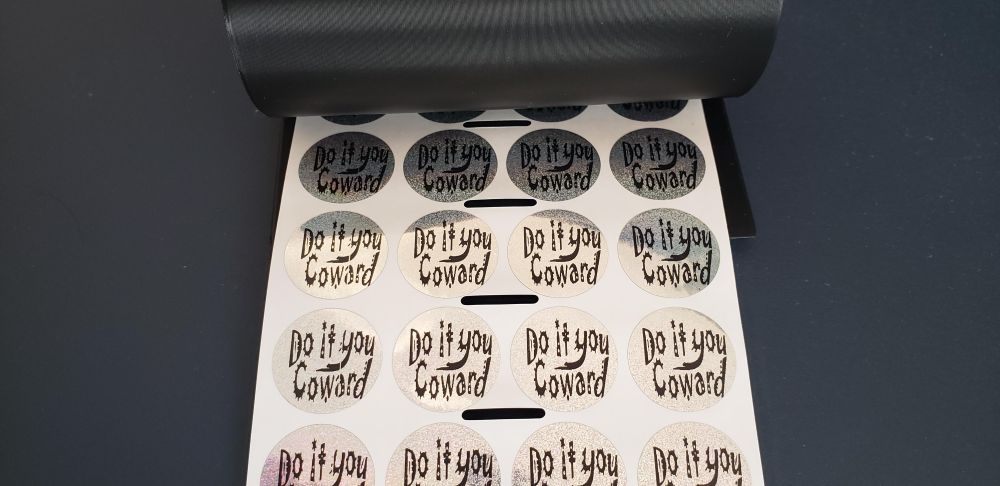 A roll of stickers in a black plastic dispenser. Each sticker is a silver holographic circle, about 1 inch diameter, with "Do it you Coward" written in the Jokerman typeface.
