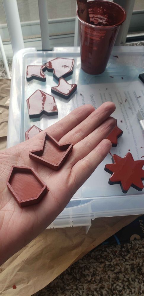 A photo of a hand holding two brick-red rubber molds, about 5mm deep and 40mm across, in two geometric shapes common to Moroccan zellij tiles. In the background are more rubber molds, still encased in the black plastic forms that they were poured in.