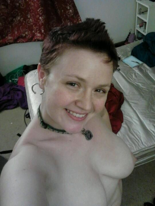 A selfie of a femme Minx from 2014, wherein they are 25, long before they started testosterone. They are smiling up at the viewer and appear fully nude, their left boob and bare tummy visible. They have a red faux hawk, and their chest lacks their distinctive Star Trek tattoo. The background of the image is littered with cords, fabric, and sex toys. 