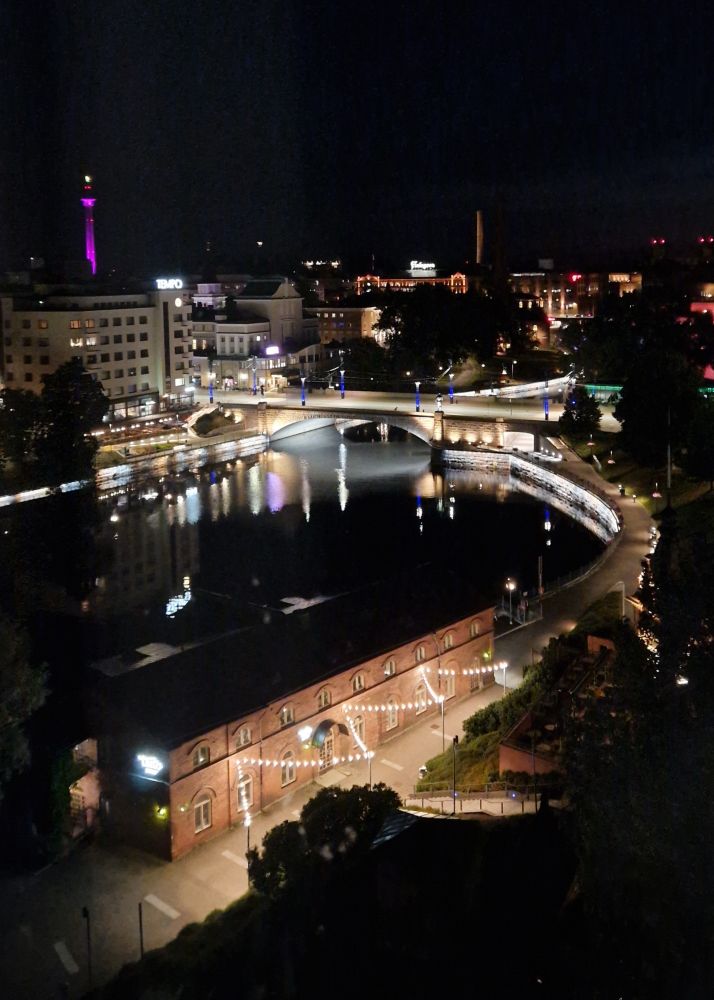 nighttime view of tampere city centre from high above. it's very dark. tammerkoski rapids with surrounding buildings, hämeensilta bridge over the rapids and näsinneula observation tower in the back, lit up in violet. it's summer.