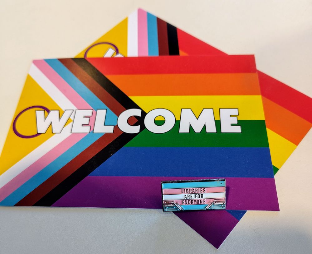 A couple of postcards with the Progress Pride flag (rainbow across and a tringle in the side with black and brown stripes followed by the trans pride colours, with the intersex symbol in the center) on top of that is a pin badge reading 'Libraries are for everyone' above the trans pride flag