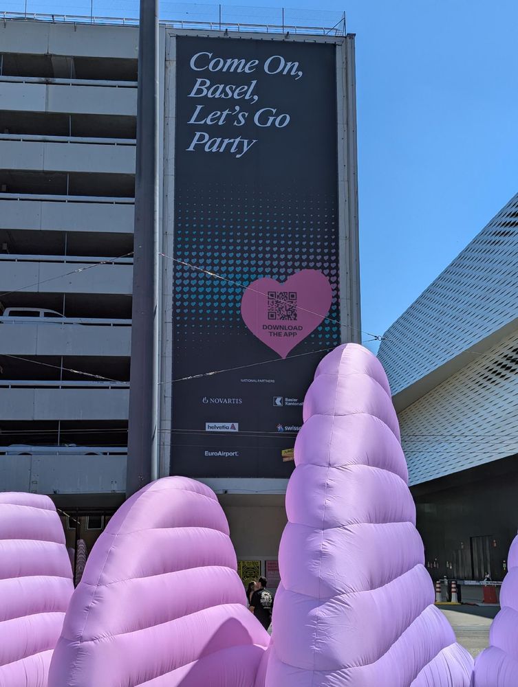 Messeplatz Basel, a colourful art installation in the foreground, a tall building in the background carrying a sign stretching along the height of the building, reading "come on, Basel! Let's party!" and displaying a stylised heart, the logo of the Eurovision Song Contest in Basel