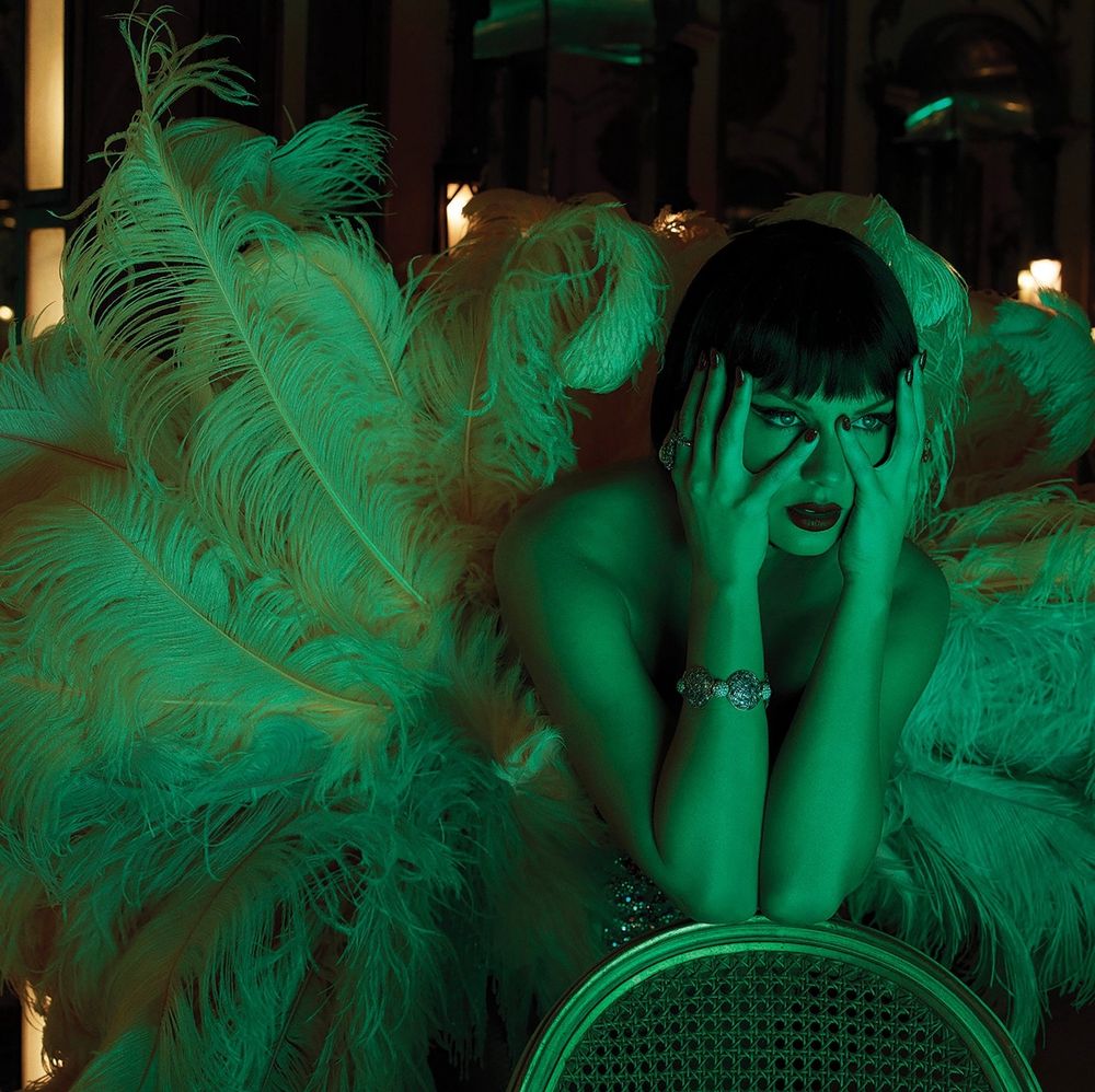 Taylor Swift in a dimly lit room with green lighting. She is wearing the same feather outfit from photo 1 but is leaning on a chair with her hands in her face 