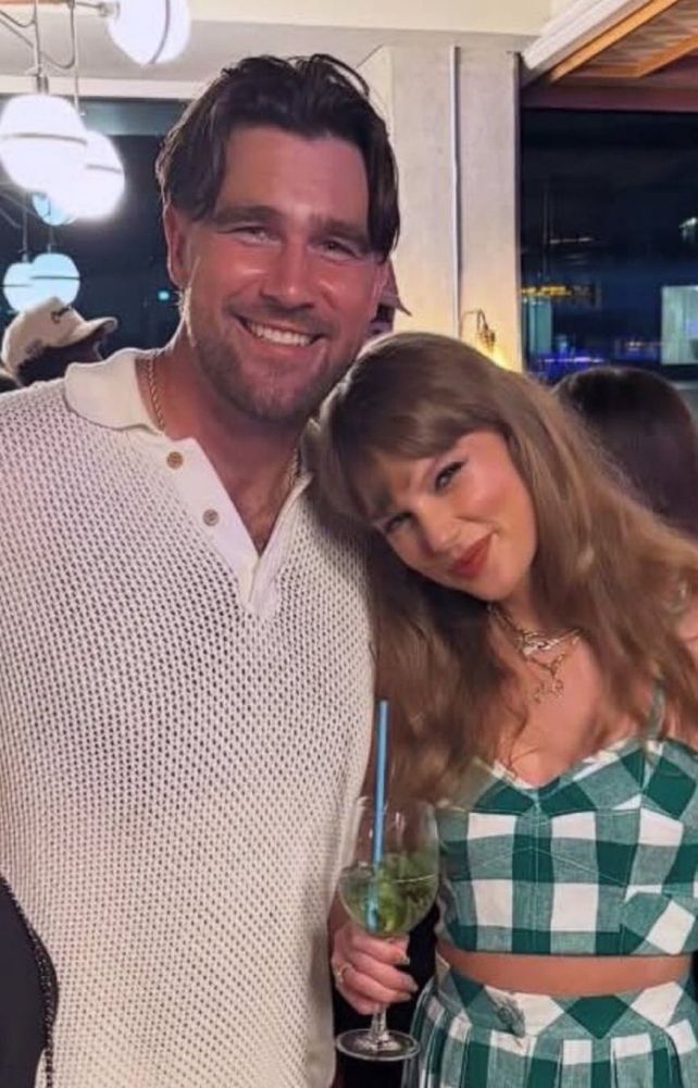 Travis kelce on the left in a white shirt. Taylor Swift in the right holding a drink in a Greek checked crop top/skirt set