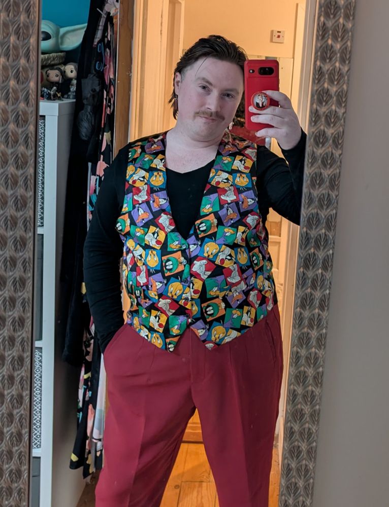 Picture of Tadhg. They are wearing a long sleeve black top, maroon trousers, and a waist coat with Looney Toons characters on it.