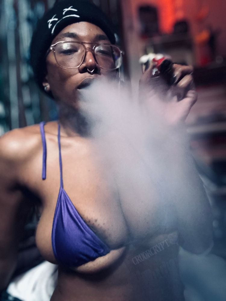 Vampyrx smokes from their severed finger chillum in their purple bikini, a peek of their nipple is seen through a haze of smoke. 