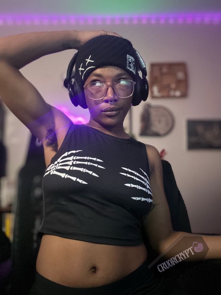 Vampyrx sits in their computer chair with a hand above their head. They’re wearing a black graphic beanie with a sad face and x’s for eyes, a black cropped tank with skeleton hands across the breast. They’re showing armpit hair as a tease. And they’re giving mad side eye. Their headphones have tape on one side.