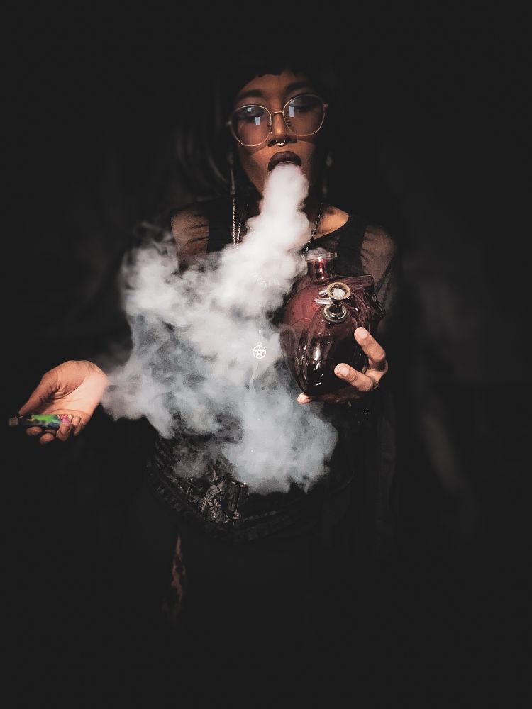 Vampyrx stands in a dark room with darkness all around them. They’re wearing black lace goal with a corset and black short bob. They have glasses on and a glasses chain dangles from the sides of their face. In their hands is a lighter and their anatomical heart bong. They exhale a large cloud of smoke from their mouth that extends down to the floor.