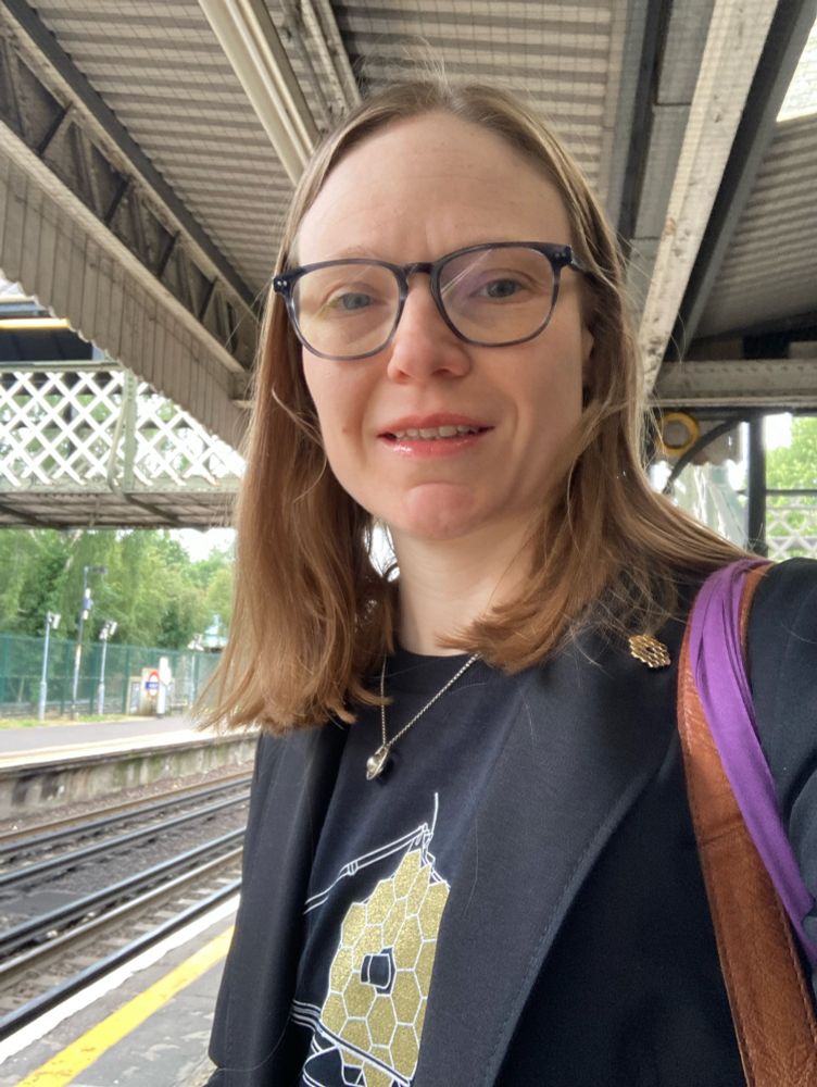 Image shows me at a tube station wearing a black t-shirt with an image of JWST on it, a black jacket with a gold coloured JWST mirror lapel pin, and a pendant necklace that looks like Saturn.