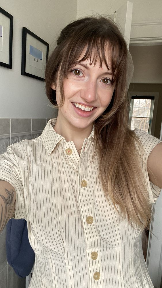 Girl with brunette hair drawn to one side of her head wearing a cream button down dress with thin navy stripes in a selfie-style picture.