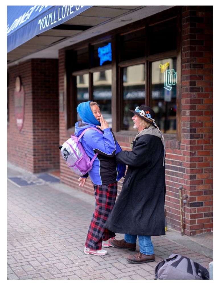 A photograph of two strangers who asked me to take their picture—the person on the left wears a periwinkle coat and flannel pajama bottoms, with a silver and pink backpack, in a pose mimicking James Bond. They are blowing imaginary smoke from the 'barrel' of their finger-guns. On the right, an individual wearing a black woolen overcoat and worn leather boots laughs at his friend's pose. He wears a wide trimmed felt hat adorned with a braided hat band of flowers that trails down his back.