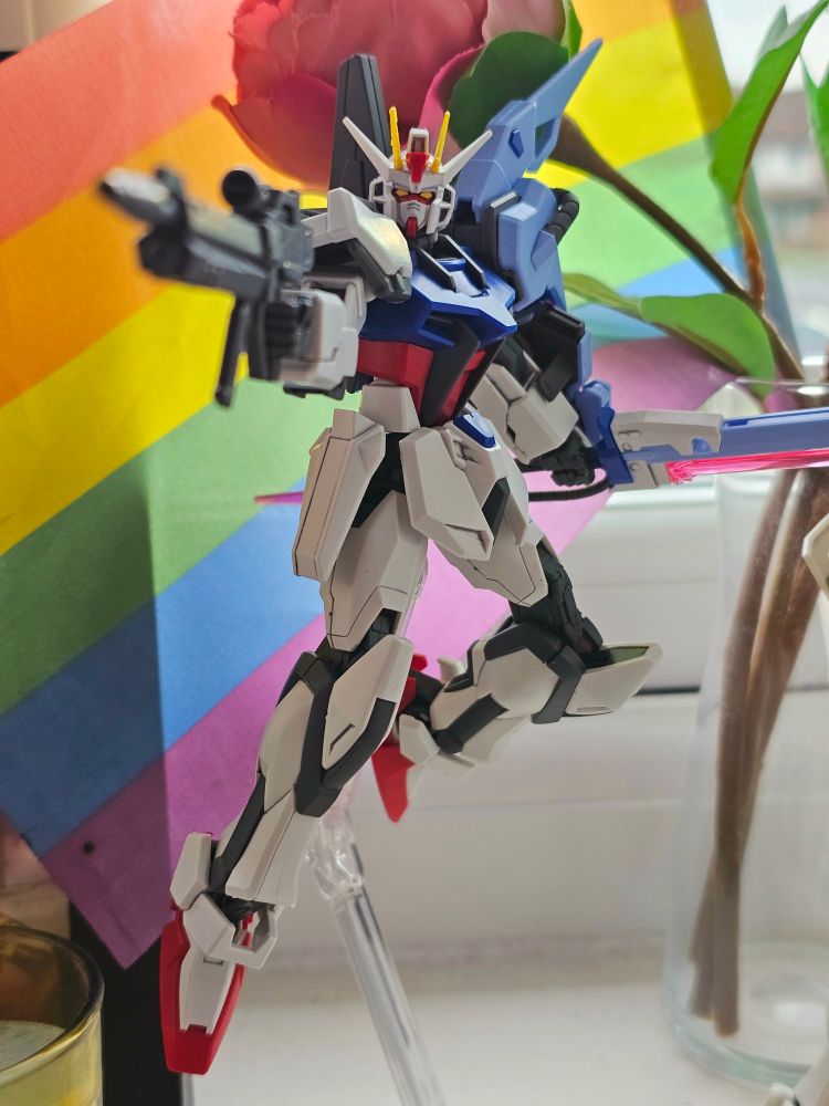 Entry Grade Strike Gundam from Gundam SEED plastic model kit on an Action Base 7 stand in front of a gay pride flag and artificial roses.
