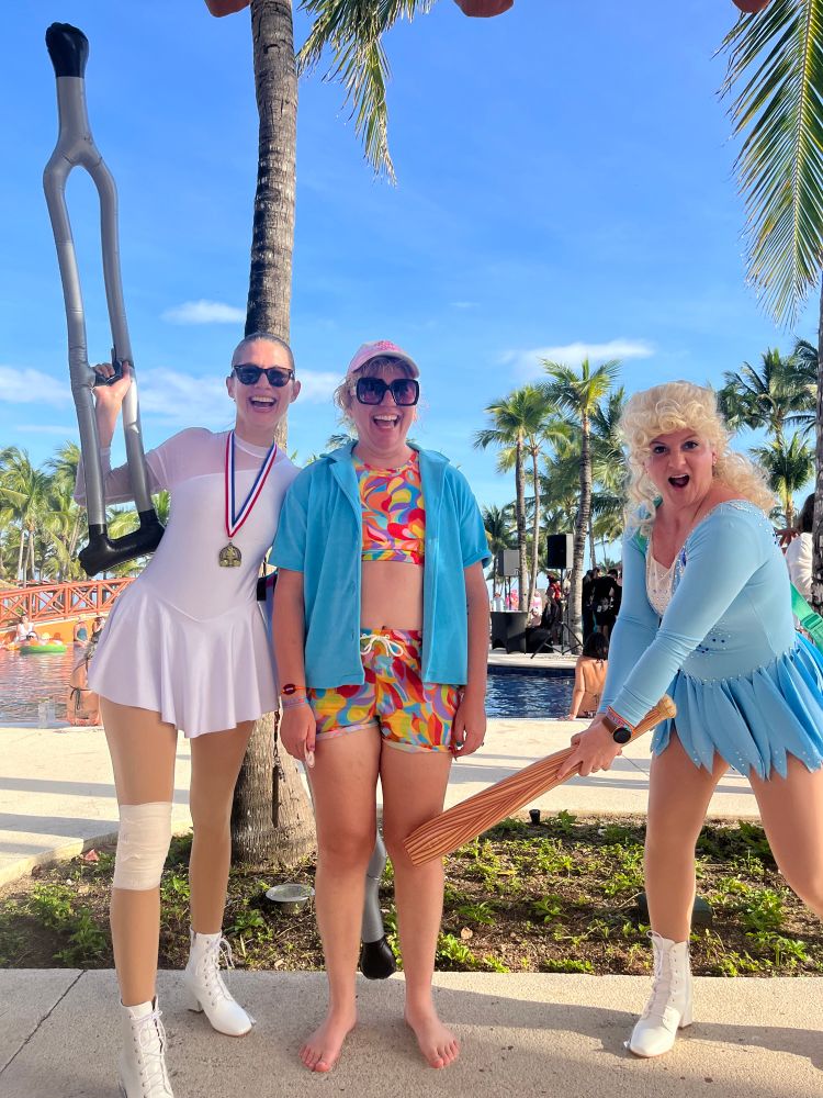 From left to right: “Nancy Kerrigan” wearing her classic white figure skating dress, a medal, knee bandage and sunglasses, holding a crutch in one hand. Linds, wearing a colorful swim suit, blue shirt and sunglasses, smiling. “Tonya Harding” wearing Tonya’s classic blue figure skating dress, a big curly wig, clubbing Linds directly in the knee. 