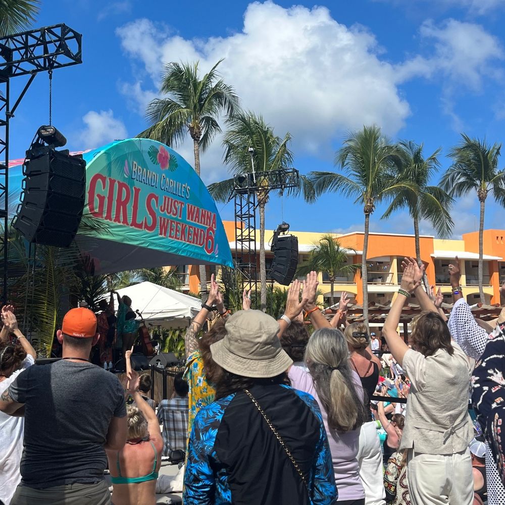 Several people with their backs to the camera, many raising their hands to clap at the performer on the stage in the background at the poolside stage, which has a big banner on top reading “Brandi Carlile’s Girls Just Wanna Weekend 6.” Brandi Carlile is in the right corner, wearing a beige vest and matching pants, clapping with her hands up. 