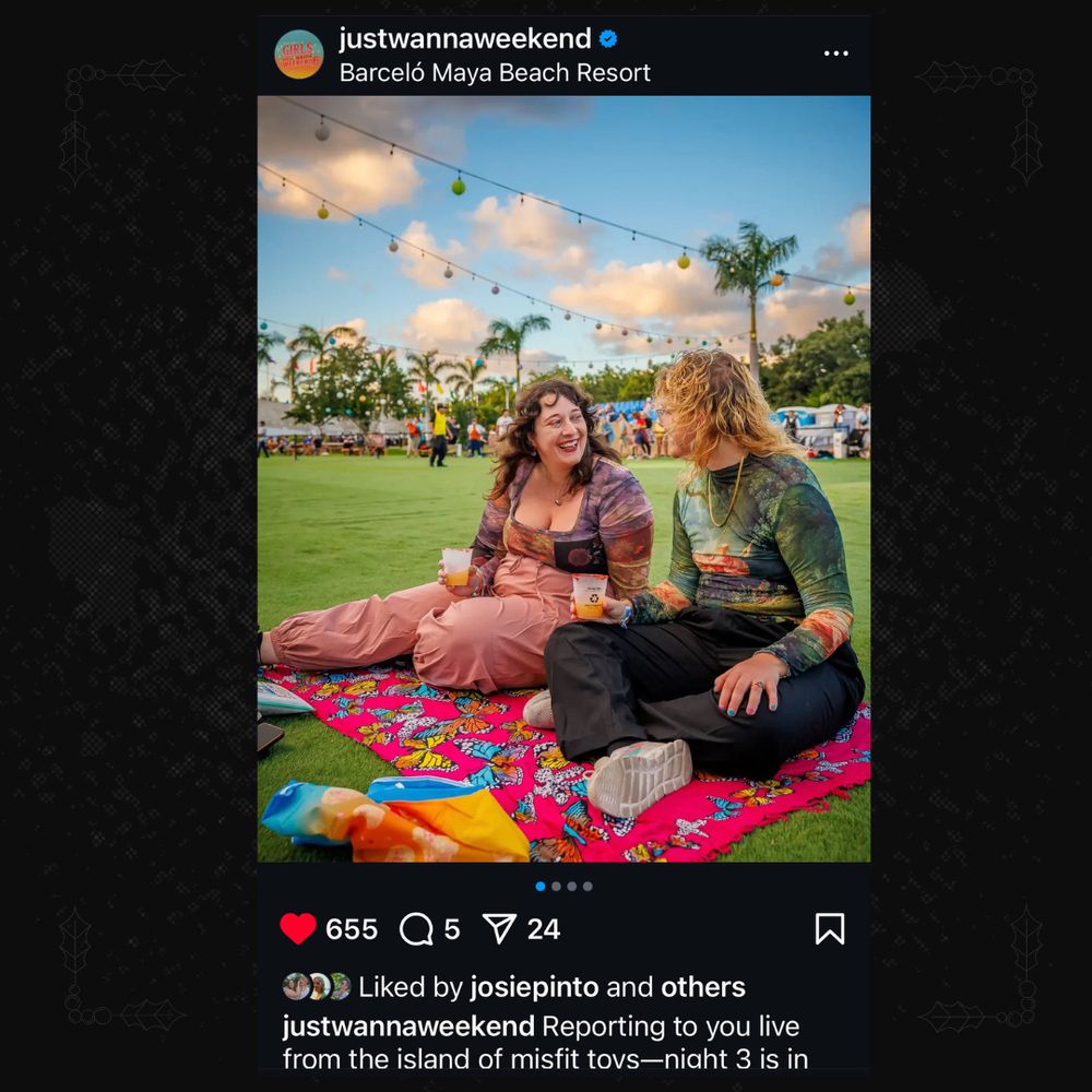 A screenshot from the official @justwannaweekend Instagram, with a photo of Josie and Linds smiling at each other and sitting on a pink throw with colorful butterflies in it, on a grass field with colorful lights and palm trees in the background, in daytime lighting. 