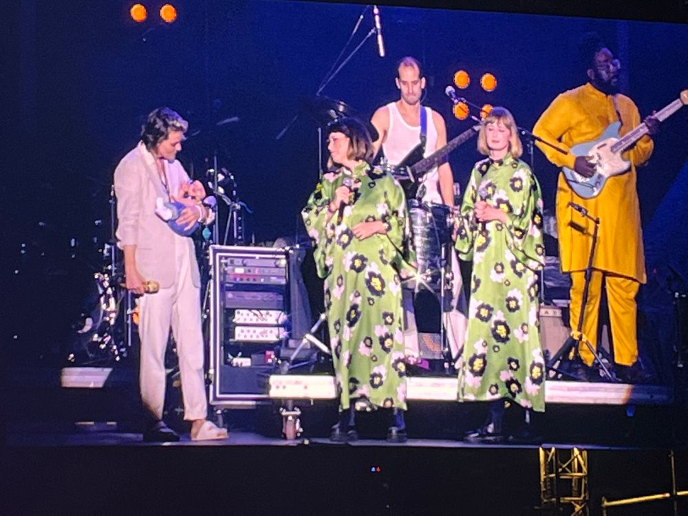 Brandi (left) wearing a beige suit, holds a small sleeping baby wearing giant headphones on stage with Jess (middle) and Holly (right) from Lucius, on stage, who are wearing long loose light green dresses with white black and yellow spots on them. 