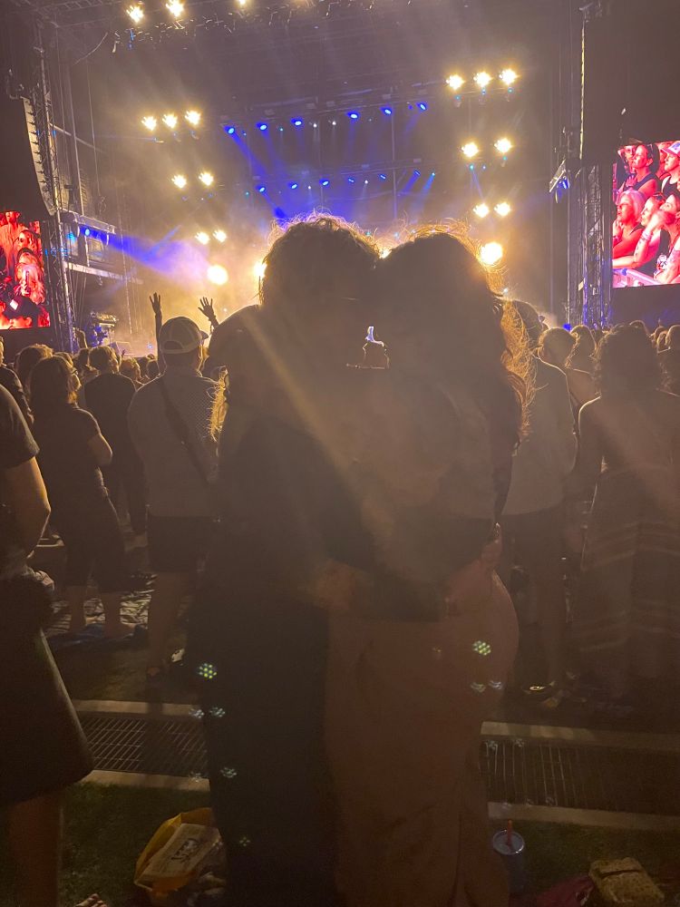 Linds and Josie hold each other close and their heads make the shape of a heart. A crowd is behind them, looking the opposite way at the stage behind them, which is bathing the dark crowd in yellow light. To the sides are part of two large screens. 