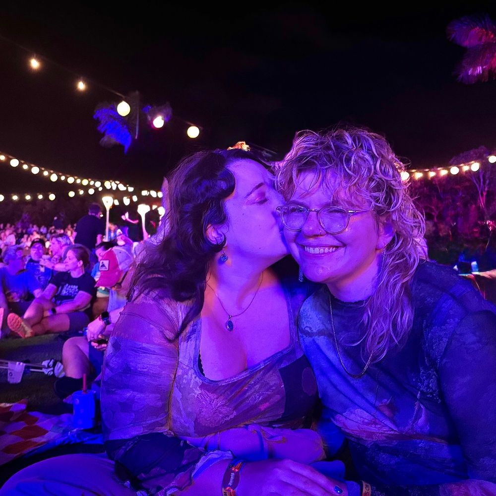 Josie, a white brunette, kisses Linds, a white person with wavy blonde hair and glasses, on the cheek while Linds smiles. A crowd is behind them and all are bathed in pinkish-purple light 
