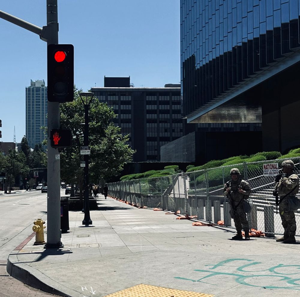 two soldiers of the national guard stand in front of a federal building on a quiet and empty street in downtown los angeles. “fuck ice” is written on the sidewalk in front of them 