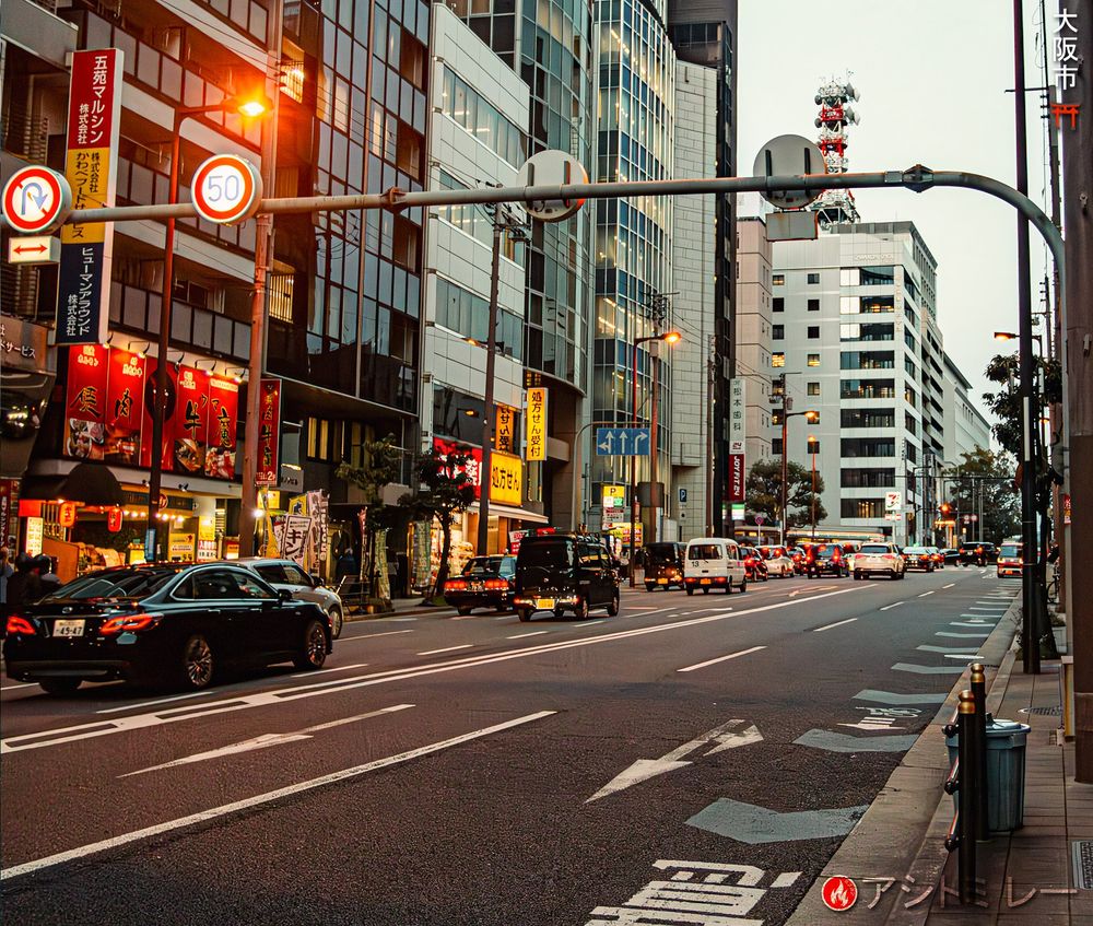 Sunset light illuminates the left of a crowded Osaka street while the right side stays blue‑gray. Shop signs glow in the dusky golden haze. | 大阪の商店街：左側は夕陽の温かな金色、右側は静かな青灰色。ほのかな灯りが通りを優しく照らす。