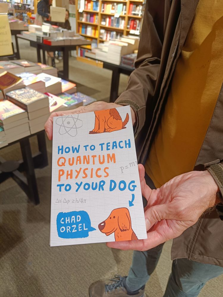 Someone (who is standing in a book shop) is holding up a book titled 'How to teach Quantum Physics to your dog'