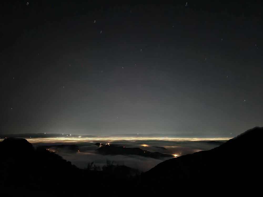 A photograph of a city as seen from the mountains above it at night. The buildings are blanketed by a low-hanging cloud layer that glows from the trapped city lights below. Above the distant glowing cloud layer, a starry moonless sky twinkles with stars.