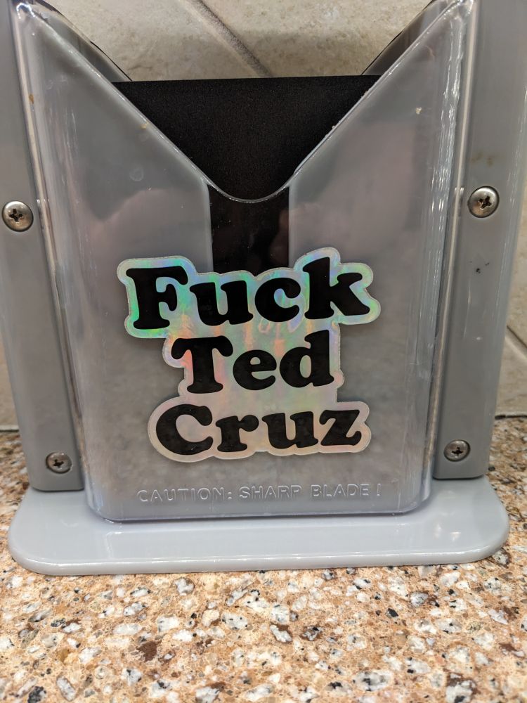 Holographic sticker on the bagel slicer saying Fuck Ted Cruz in a font like Cooper Black 