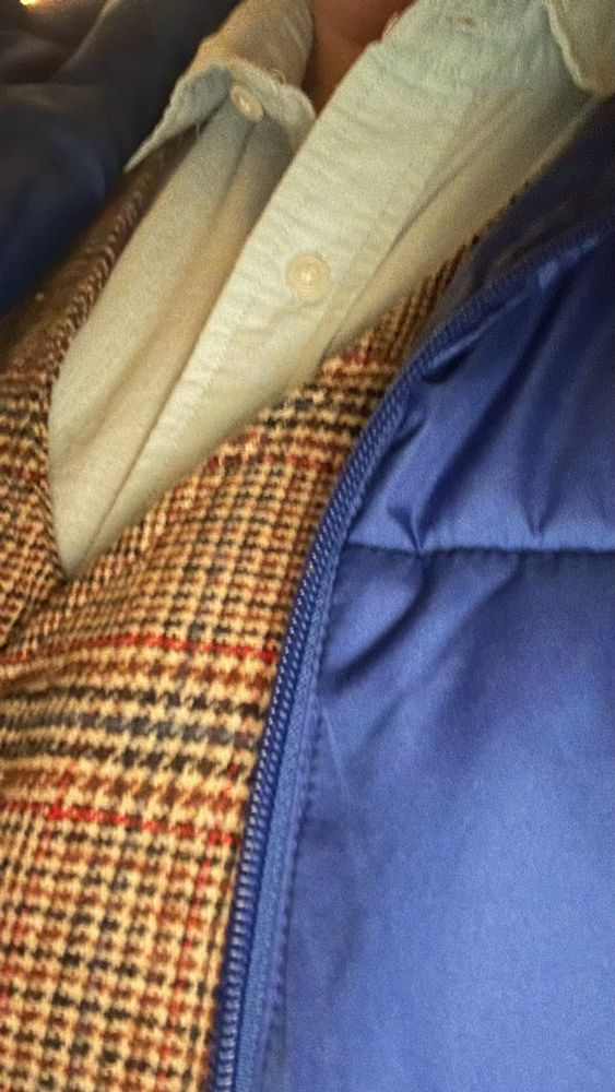 A photo showing a light blue button up shirt under plaid vest and blue puffy coat