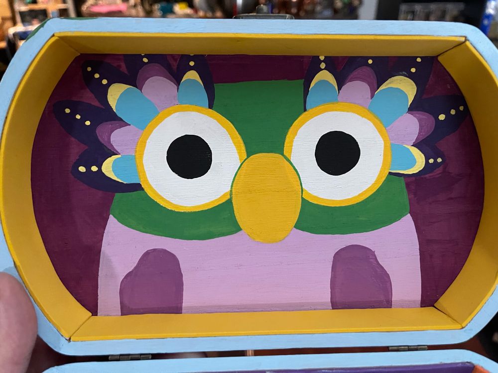 Inside the lid of a wooden jewelry box is a handpainted Chattermax from the children’s TV show Bluey, it is painted in purples and greens and oranges and light blues and yellows. The box lid is lined on the sides with golden yellow craft foam strips