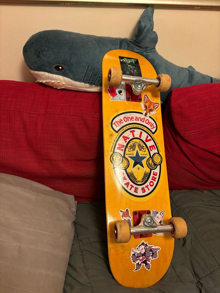 Only’s skateboard, a yellow Native Skate Store deck with a Newcastle Brown Ale inspired design, decorated with several furry stickers. It is being modelled by Blåhaj