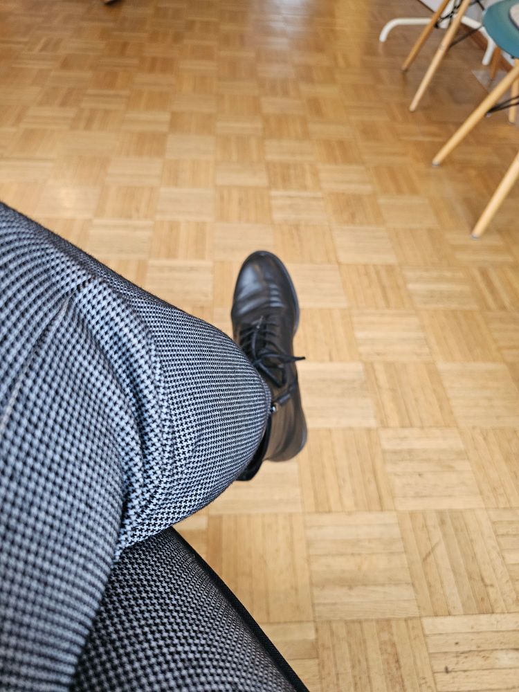  Überkreuzte Beine in kleinkarierter Hose, schwarze Stiefeletten, heller Parkettboden