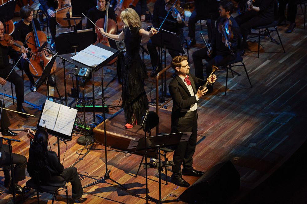 A man in a tuxedo with tails and a comically large red bow tie stands in front of a symphony orchestra on stage at Hamer Hall in Narrm, Australia. The man (me) holds an old car horn, which he is about to honk.