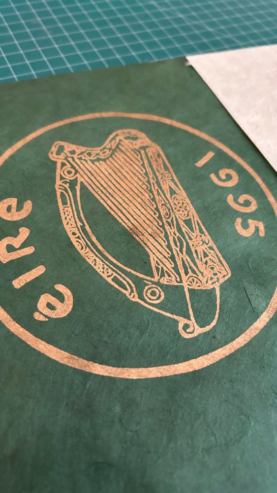 Print depicting an old Irish coin with a harp, éire and the year 1995, in gold ink on green paper 