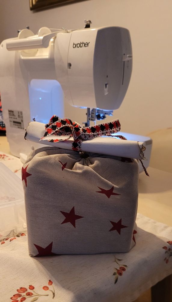 Ein genähtes Geschenksäckchen aus beigen Baumwollstoff mit roten Sternen drauf. Oben eine Schleife aus bunten Bändern und einem eingerollt Papier