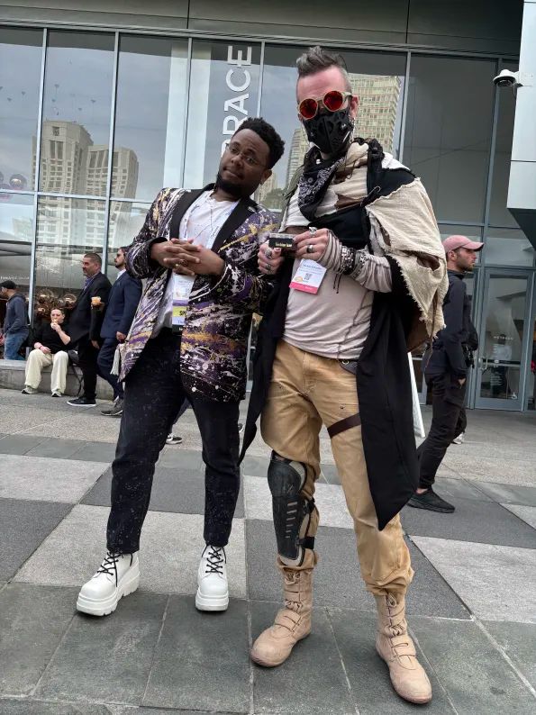 Two people posing confidently outside a contemporary building. One wears a vibrant purple and gold-patterned jacket, black pants, and white boots, leaning slightly with hands clasped. The other sports a distinctive, futuristic costume with a mask, red goggles, layered clothes, and a leg brace.