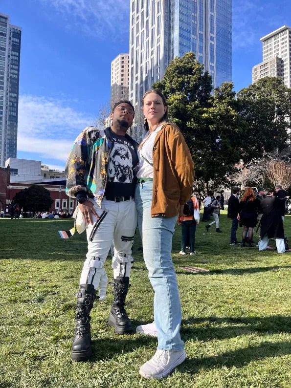 Two people standing confidently on a grassy park area in front of tall city buildings. One wears a colorful jacket, graphic shirt, white pants, and tall black boots. The other wears a brown jacket, white shirt, jeans, and sneakers, both looking proudly into the camera under a bright blue sky.