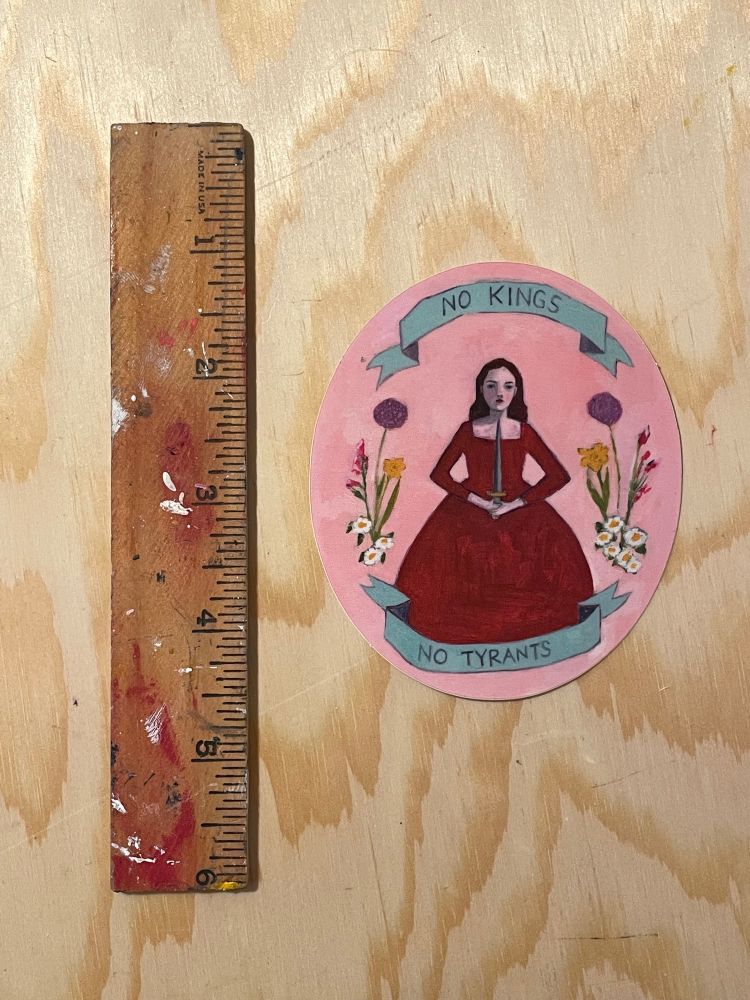 Sticker next to a ruler - image of oil painting, woman holding a sword, surrounded by flowers and banners that say “no kings” “no tyrants”