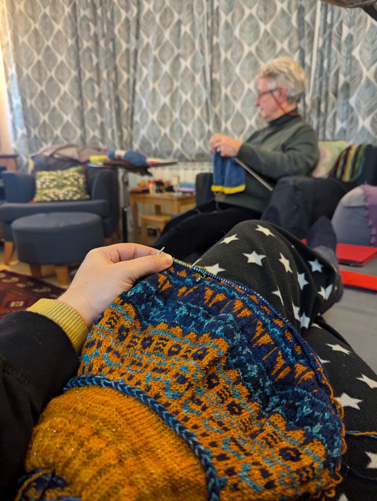 A piece of hand knitting in progress is shown in the foreground, on the photographer's lap. In the background, a white European lady in her 70s is also knitting, facing away from the camera (she is watching a David Attenborough documentary)