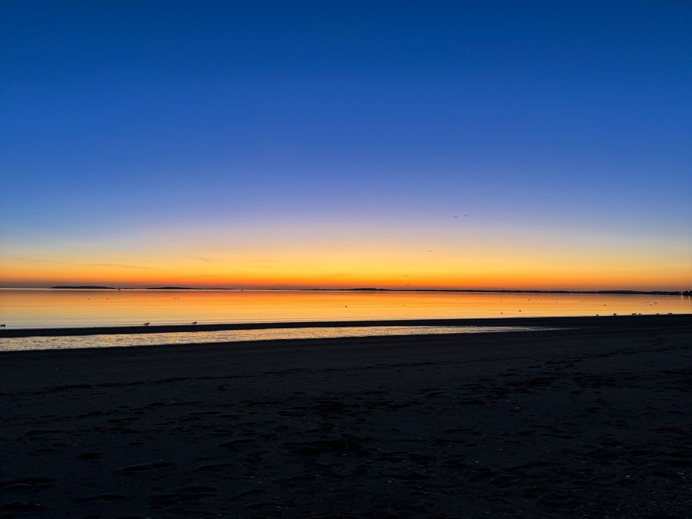 Twilight over a smooth sea & beach cloaked in darkness, with a narrow glowing-ember horizon streaking through the dark indigo sky.