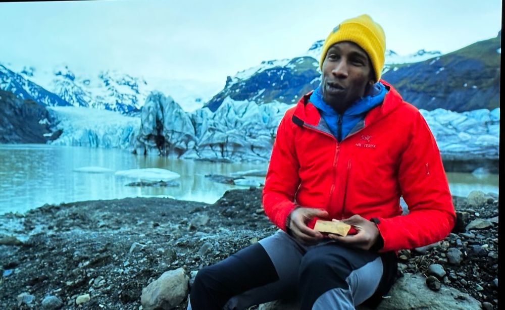 A Black man (Chris) in red jacket & yellow hat sitting in front of a rapidly advancing glacier with a rock in his hand.