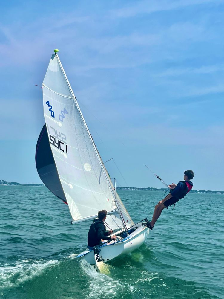 Two teens in a small 420 sailboat out sailing on the ocean with the spinnaker sail out . The sailboat is tilted about 45 degrees & one teen is in a trapeze harness hanging all the way out over the edge of the boat to balance the force of wind in the sails & keep boat from tipping over. 