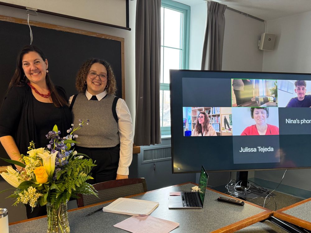 Alisa & Leticia smiling at successful conclusion of defense with a vase flowers & zoom screen with some remote audience & committee members. 