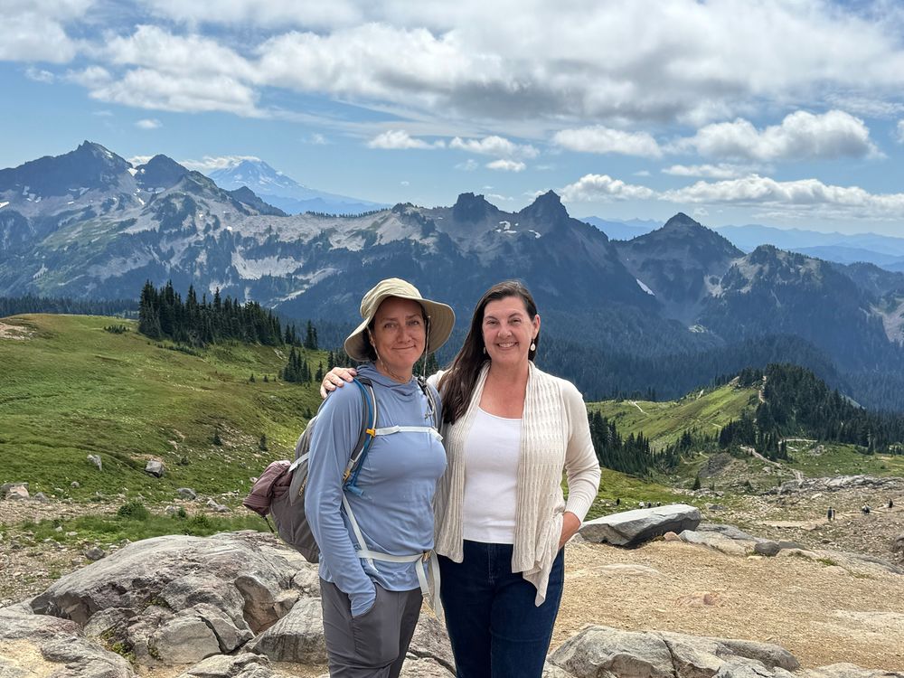 Two women up on a high vista point with mountains behind them. 