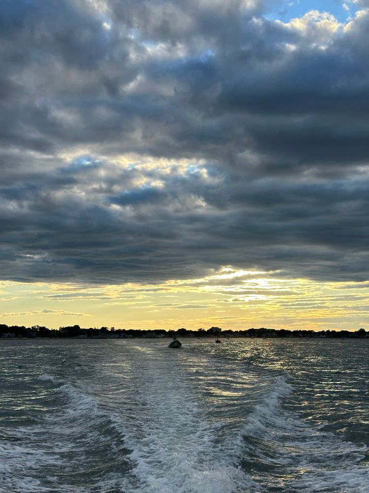 Dramatic sky with gray brooding clouds over head & reflected in a turbulent gray sea with a wake, & a band of golden yellow sunset glowing between the gray sky & sea. 